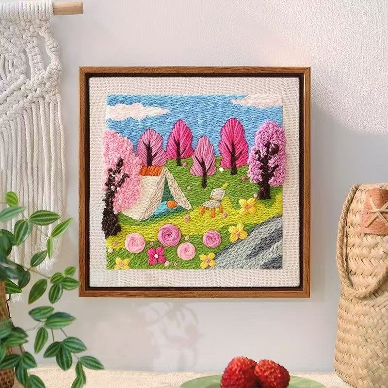 Framed yarn embroidered artwork of a campsite with trees and flowers on a wall.