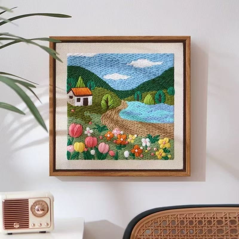 Framed yarn embroidered landscape with a house, trees, and flowers on a white wall.