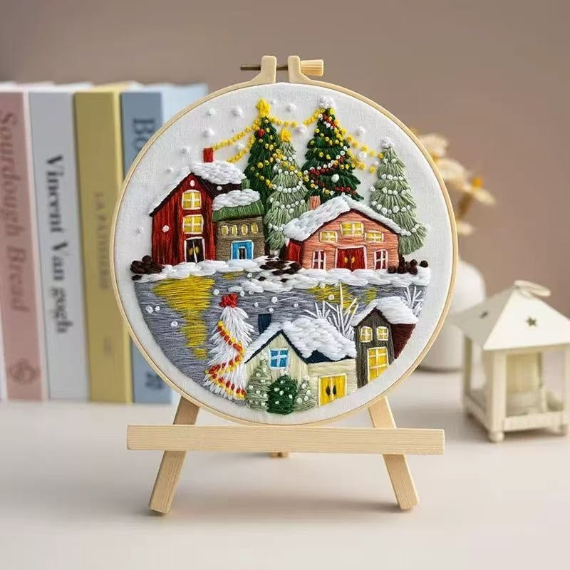 Embroidery of a snowy village scene on a wooden hoop with books in the background.