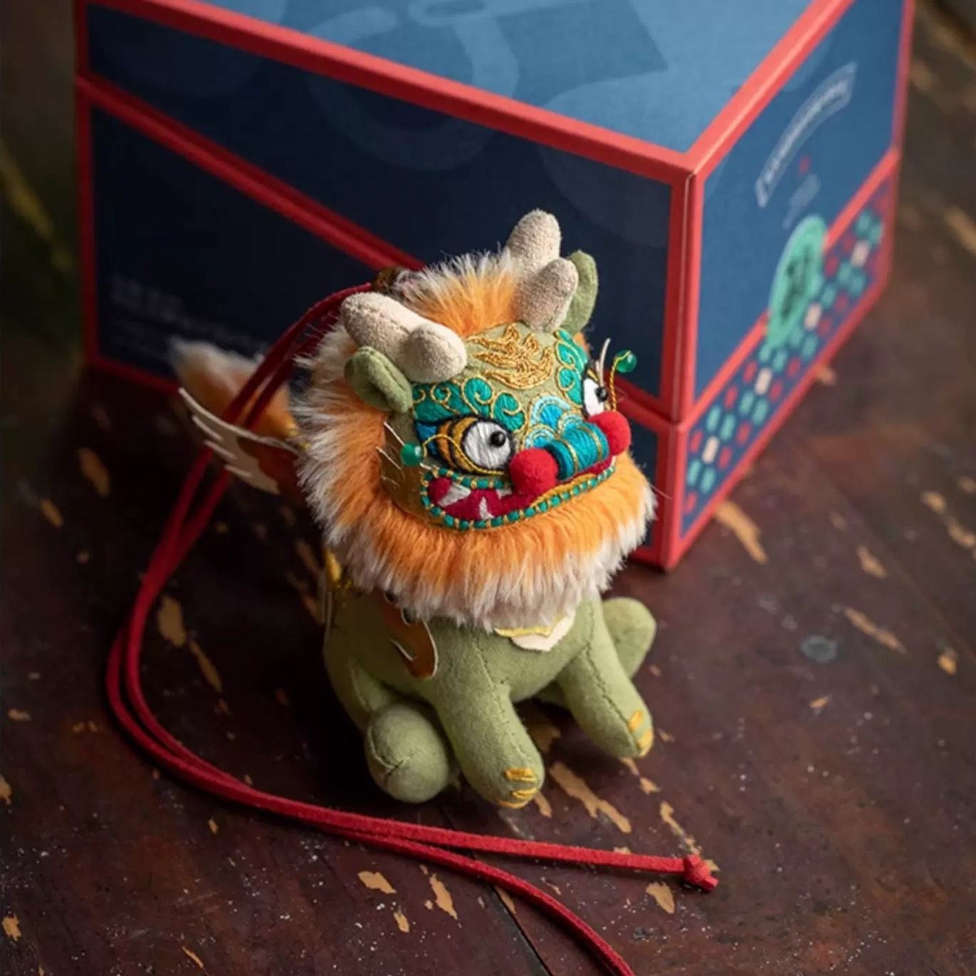 Stuffed Chinese beast Kylin toy doll with colorful face on a wooden surface, with a gift box in the background.