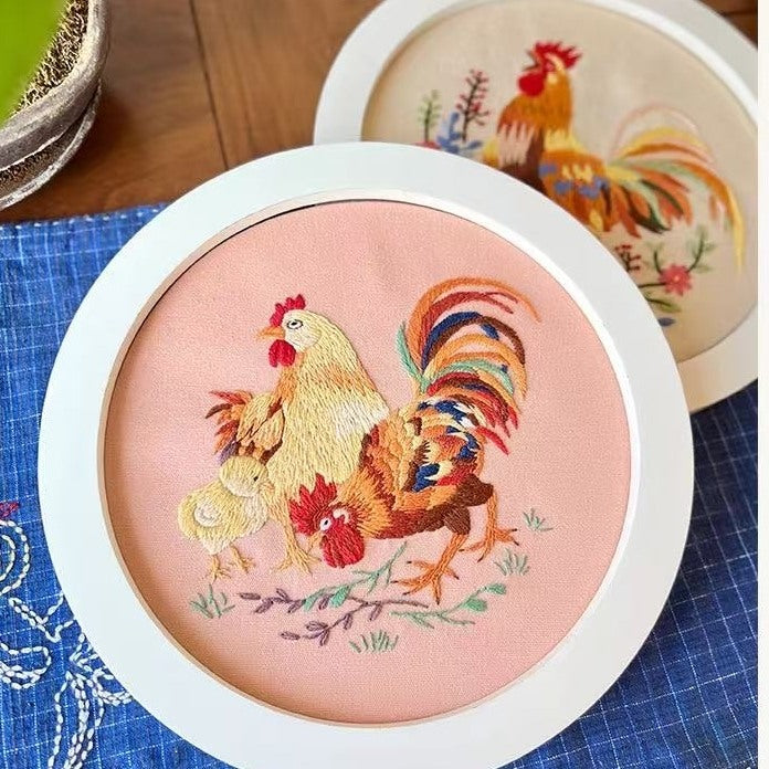 Embroidery hoop with rooster design on a pink background, placed on a blue fabric surface.
