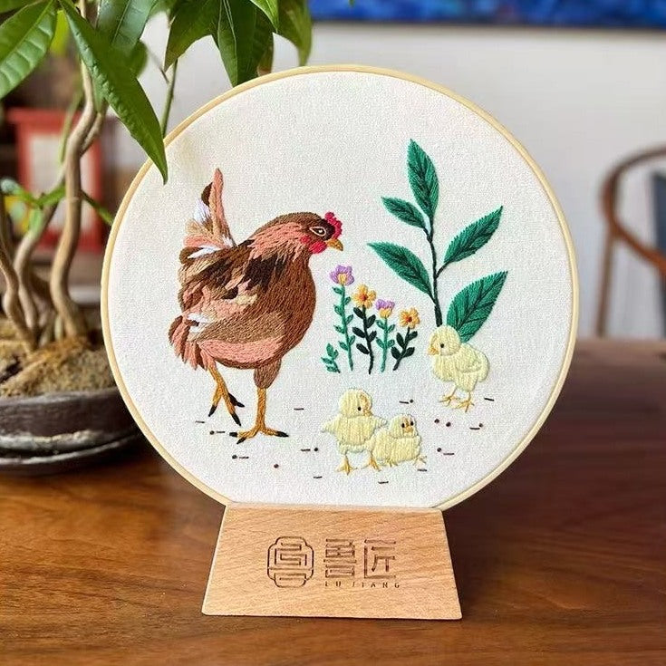 Embroidery of a rooster and chicks on a circular wooden stand with a plant in the background.