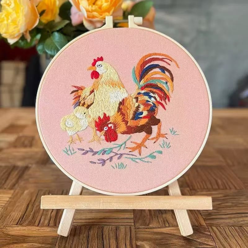 Embroidery hoop with rooster and chicken design on a pink background, placed on a wooden surface.