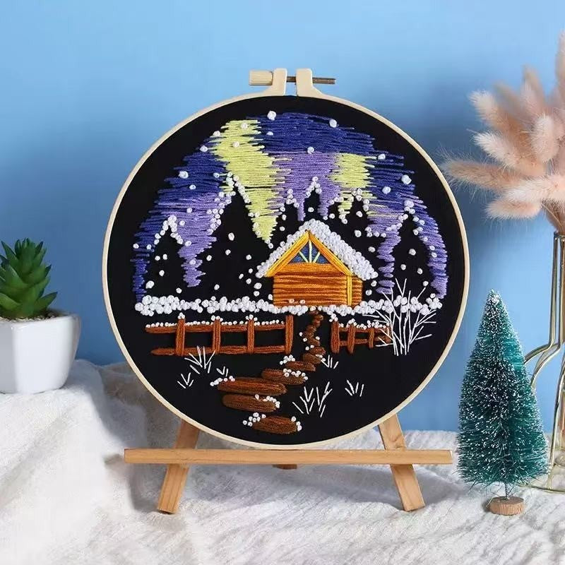 Embroidery hoop with a winter scene design featuring a cabin, trees, and snow on a blue background.