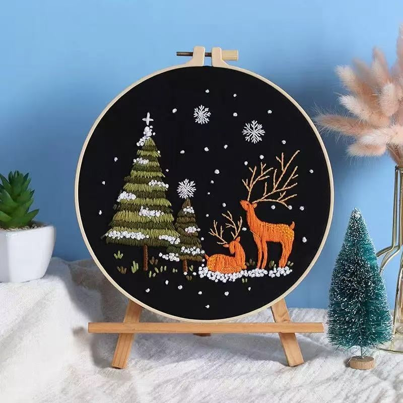 Embroidery hoop with deer and Christmas tree design on a stand against a blue wall.